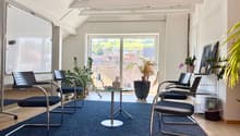 Spacious office space with large windows overlooking a scenic view, modern furniture including black chairs and a glass-top table, and various potted plants decorating the room.