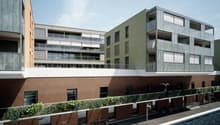 Multi-story apartment building with a modern, minimalist design. The building features a mix of materials including wood, glass, and brick. There are balconies on some of the units, providing outdoor space for the residents. The building appears to be par