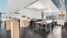 Large open-plan office space with high ceilings, white walls, and concrete flooring. The space contains various workstations, desks, and chairs, as well as architectural models and displays on shelves. There is a view of the city through the large windows