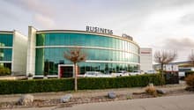 A large, multi-story glass and concrete building with the text 'BUSINESS CENTER' prominently displayed. The building has a curved facade and is surrounded by a parking lot with several cars parked. There is landscaping with trees and shrubs around the bui
