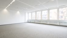 Large open office space with multiple windows providing a view, white walls, gray flooring, suspended ceiling with recessed lighting