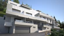 3-story modern apartment building with a gray and white exterior, balconies, and a garage on the ground floor. The building is surrounded by trees and has a sloped driveway leading up to the entrance.