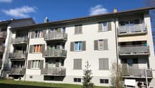 White apartment building with multiple balconies, brown roof, multiple windows, green grass in front