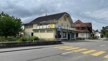 A two-story commercial building with a yellow exterior, a tiled roof, and several windows. The building has a grocery store called 'Swissco' on the ground floor. There is a crosswalk in front of the building and the surrounding area appears to be a reside