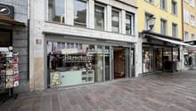 The storefront is a multi-story building with large windows, glass doors, and awnings. Inside the storefront, there is a shop with a display of products. The storefront is located in a busy street with other shops and a brick sidewalk.