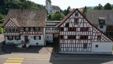 Building with a traditional design, two stories, brown and white checkered exterior, large windows, entrance with a staircase, sign reading 'Wurschaus Sormen', small wooden bench, church with a steeple in the background, green surroundings