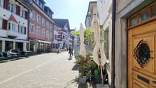a cobblestone street, people walking, TAKE AWAY sign, outdoor dining tables, potted plants