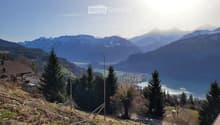 Aerial view of mountainous terrain with lush vegetation, a serene body of water, distant houses, and a clear sky.