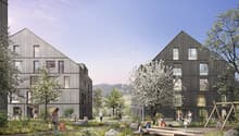 The image shows a residential complex with several multi-story wooden buildings surrounded by a lush, landscaped outdoor area. The buildings have balconies and large windows, and the outdoor space features trees, flowers, and a play area with logs and ben