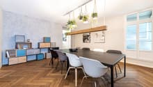 The image shows a modern and stylish dining room. The room has a large wooden dining table with black chairs around it. Above the table, there is a wooden beam with hanging plants in macrame planters, adding a touch of greenery to the space. The walls are