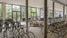 large room with many bicycles parked, wooden floor, large windows, metal poles, tables with bags