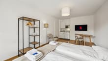 Compact bedroom with wooden floor, double bed, microwave, tv, dining table, small kitchen, hanging cabinet, floor lamp, and chair.