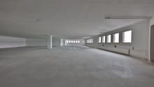 Large open space with concrete floor, white walls, and multiple windows along the sides. The space appears to be an unfinished commercial or industrial building, possibly a warehouse or factory.