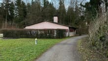 Pink building with a brown roof, pathway leading up to the entrance, hedge on the side, and a grassy field in the background.