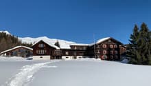 A wooden building covered in snow with several windows, two smaller houses on the left, trees in the front, mountains in the background