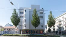 A multi-story commercial building with a modern, minimalist design. The building has large windows and a flat roof. There are several trees lining the street in front of the building. The ground floor appears to house a retail or restaurant space with sig