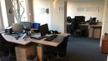 The image shows an office space with multiple desks, computers, and office equipment. The room has large windows providing natural light, and the walls are painted a light color. There are various office supplies and documents visible on the desks.