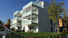 Modern multi-story apartment building with balconies, glass windows, and a landscaped yard with trees and shrubs.