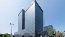 Large modern glass and steel office building with a rectangular, geometric design. The building has multiple floors and a prominent facade with a grid-like pattern of windows. There are trees and other buildings visible in the surrounding area.
