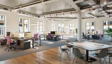 Large open-plan office space with high ceilings, large windows, and modern furniture. The space features workstations with computers, conference tables, and a mix of seating options. The decor is minimalist with a neutral color palette and wooden flooring