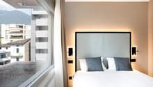 Double bed, large window with view of mountains, white walls, two bedside lights, gray headboard, white sheets, gray pillows