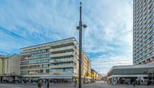 The image shows a busy city street with a mix of commercial and residential buildings. There is a tall lamp post with multiple cameras mounted on it in the center of the frame. People can be seen walking along the sidewalks and crossing the street. The bu