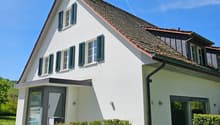 Modern white house with a brown roof, green shutters, and a glass door.