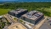 Large modern office complex with multiple interconnected buildings, surrounded by green landscapes and mountains in the background. The buildings have a sleek, contemporary design with glass facades and solar panels on the roofs. There is a parking lot in