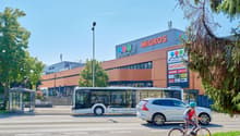 A large Migros supermarket building with a bus stop and people riding bicycles in front of it. The building has a distinctive orange and blue color scheme.