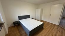 The image shows a bedroom with a double bed, a bedside table, and a wardrobe. The room has white walls and a hardwood floor.