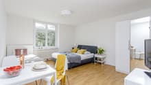 Bright and spacious studio apartment with large windows, hardwood floors, a double bed, a dining table, and a TV stand. The room has a cozy and modern feel with yellow accents and decorative elements.