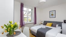 Spacious bedroom with large windows, gray upholstered bed, white bedding, purple curtains, and a vase of yellow tulips on a side table