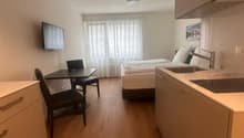Compact studio apartment with hardwood floors, white walls, a bed, a desk with a chair, and kitchen cabinets with a dishwasher visible.