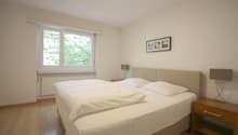 Spacious bedroom with large windows overlooking greenery, white walls, hardwood floors, a king-size bed with white bedding, a wooden nightstand, and a framed artwork on the wall.