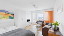 Bright and spacious studio apartment with large windows offering a view of the city. The room features a bed, a sofa, a dining table, and a TV stand. The walls are decorated with a colorful landscape painting. The apartment has hardwood floors and modern