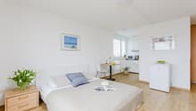 white walls, wooden floor, single bed, pillows, hat, table with flowers, kitchenette, refrigerator, picture frames