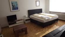 a bedroom with wooden floor, large bed, night table, TV on TV stand, a table, and a couch