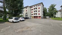4-story apartment building with white exterior and red shutters, parking lot in front of the building, surrounded by trees
