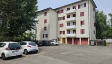 4-story apartment building with beige exterior and red shutters, parking lot in front with several cars parked