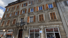 Multi-story building with a traditional European architectural style, featuring stone walls, wooden shutters, and a balcony. The building houses various commercial establishments, including a lottery shop, an art studio, and a cafeteria.