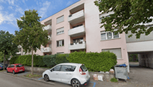 3-story apartment building with balconies, surrounded by trees and greenery, with parked cars in the foreground