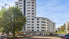 White high rise building with many windows, street tree, parked cars, shops on street level