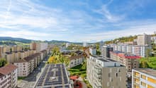 Aerial view of an urban setting with residential buildings, rooftop solar panels, greenery, and distant mountains under a clear sky