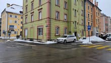 Corner building with four floors, light green color with red frames around the windows, snow on the ground, a parked car in front, two other buildings next to it