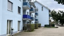 Blue apartment building with multiple balconies, brick paved driveway, hedges, trees