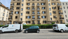 Multi-story apartment building with beige facade, balconies, and windows. Several parked cars visible in front of the building.