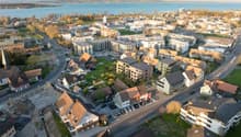 Aerial view of a city with a mix of residential and commercial buildings, surrounded by a body of water and hills in the background. The city appears to have a mix of older and newer buildings, with a variety of architectural styles. There are roads, park