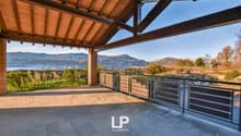 Covered outdoor terrace with wooden beams and tiled flooring, offering a scenic view of the surrounding mountains and lake in the distance.