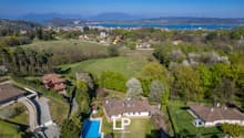 Aerial view of a large house with a swimming pool surrounded by lush greenery, with a scenic view of a lake and mountains in the background.