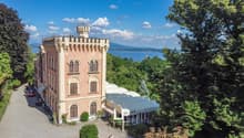 A large, pink-colored castle with turrets and arched windows, surrounded by lush greenery and overlooking a body of water in the distance.
